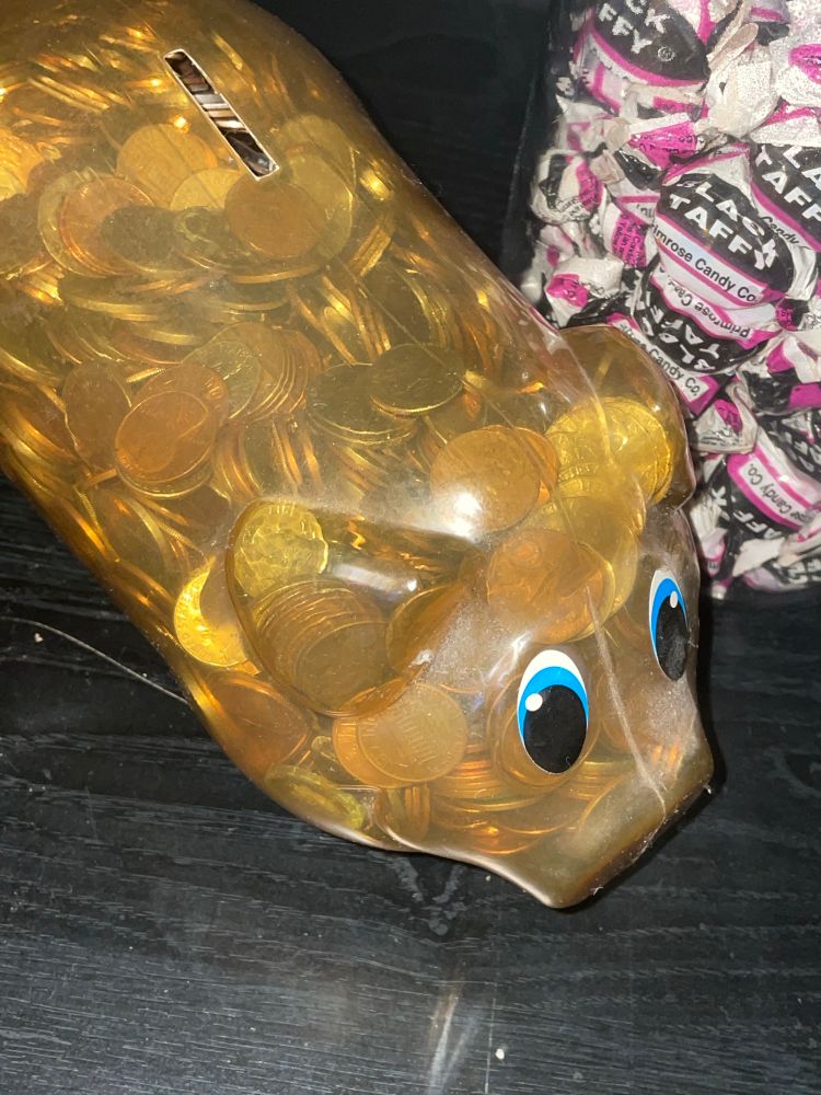 A clear plastic piggy bank sitting on a black shelf. A jar of black and pink taffy sits next to it. 
