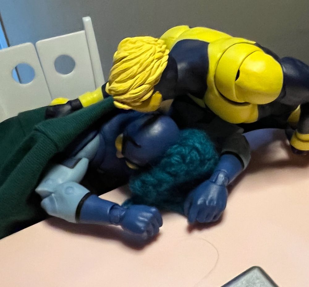 Booster gold action figure closes computer and puts Ted Kord action figures head on pillow and blanket over him and kisses his head