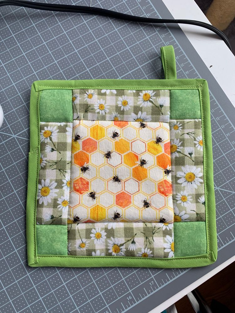 A square quilted potholder. It is mostly green with a bee/honeycomb pattern fabric and a plaid fabric with daisies on it.