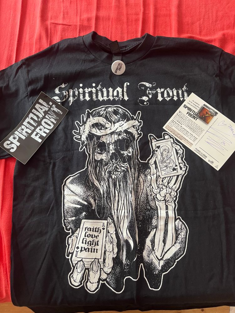 Die Vorderseite vom Shirt mit Jesus als Skelett mit langem Bart. In der rechten Hand hält er ein Kartenspiel und auf der ersten Karte steht „faith love light pain“. Die linke Hand hält er eine Karte in Kopfhöhe. Auf der ist die Jungfrau Maria zu sehen mit einem Kind im linken Arm und einer Pistole in der rechten Hand. Oben drüber steht „Spiritual Front“