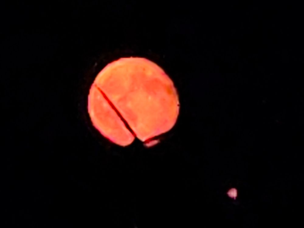 Unscharfes Handyfoto... Zoom in den Abendhimmel. Ein orange-rötlicher, abnehmender Mond steht am dunklen Himmel. Ein Flügel eines Windrades schneidet schräg durch den Mond und erzeugt einen dunklen Schatten. Unten rechts im Bild ist ein kleines, helles Licht zu sehen, das vermutlich ein weiterer Windradpunkt oder ein Licht in der Ferne ist.