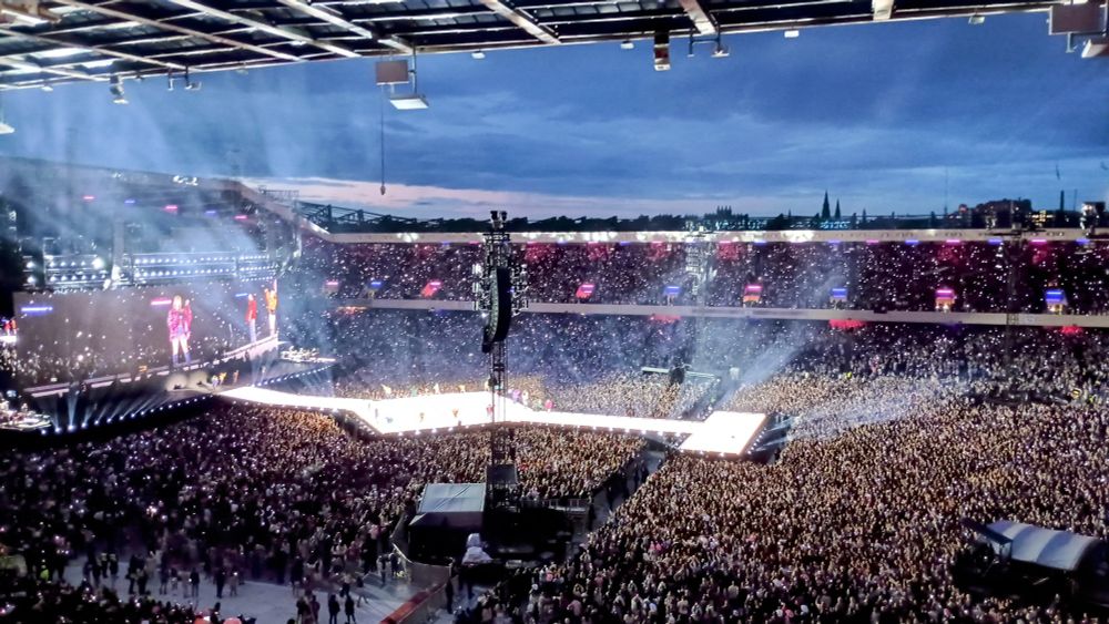 The Eras Tour in a full stadium with an open roof. The view is from the top of the stadium. Taylor Swift can be seen on the screen in the far left. The dancers are all around the diamond in the center of the stage. They are all wearing different color fringe jackets. The stage is lit up. The crowd has light up bracelets which can also be seen glowing. 