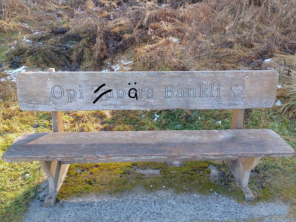 Eine hölzerne Sitzbank, in deren Rückenlehne geschnitzt steht: Opi Zubers Bänkli. Danach ein Herz-Symbol. Ich habe das Bild bearbeitet: Das "Zu" in "Zubers" ist mit zwei schwarzen Strichen durchgestrichen, über das "e" ist ein "ä" geschrieben.