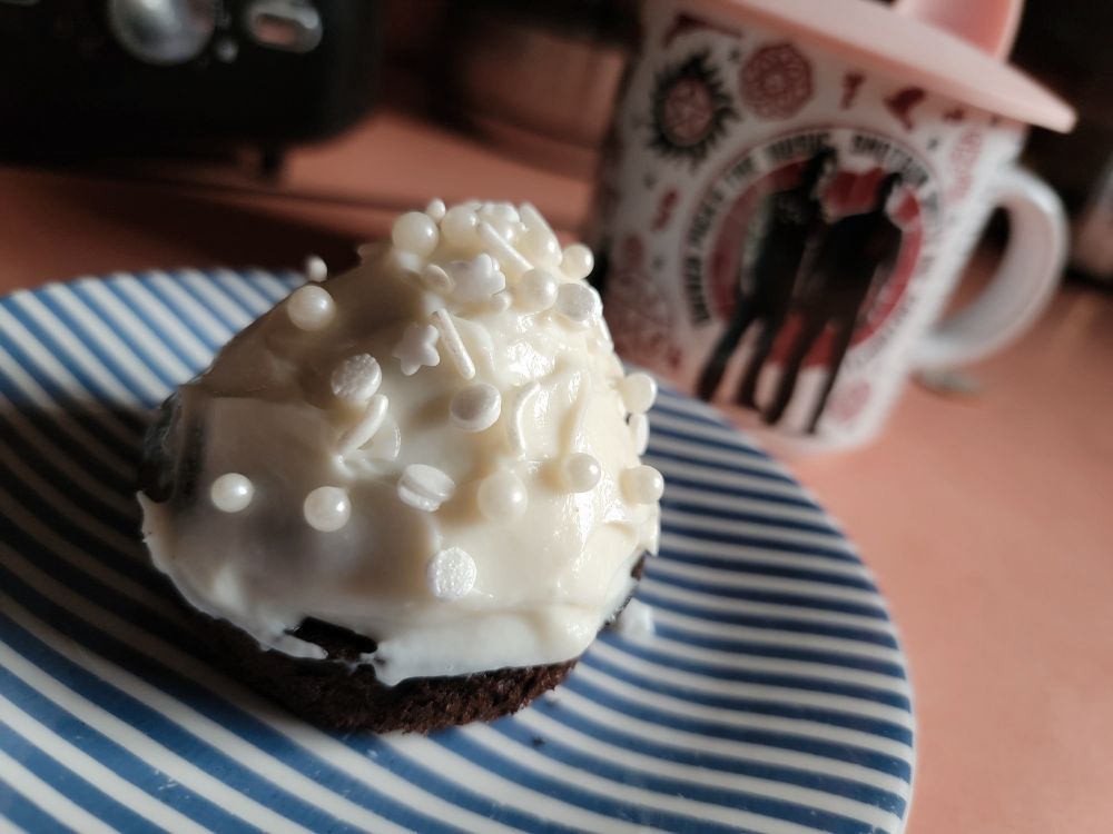 a single chocolate cupcake with cream cheese frosting and white pearl sprinkles displayed on a blue and white striped plate. in the background is a supernatural coffee cup displaying a black and red print of sam and dean from supernatural and various related symbols.