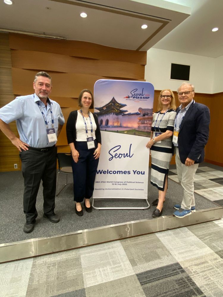 Foto von vier Personen vor einem Aufsteller des IPSA World Congress 2025 in Seoul. Von links nach rechts: Aurel Croissant, Eva Heidbreder, Marianne Kneuer und Ferdinand Müller-Rommel.