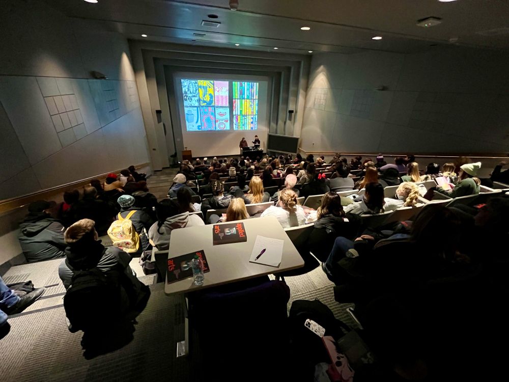 View to the podium of two speakers in front of a screen, in a lecture hall.