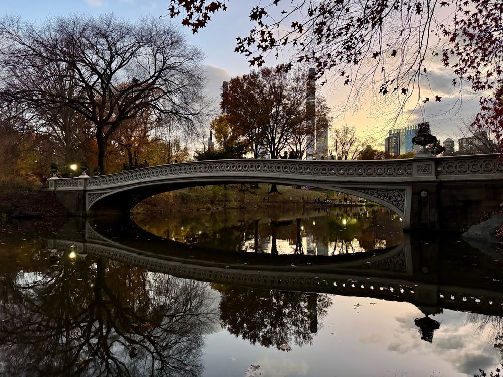 Bow Bridge across a lake in Central Park, the lake reflects the bridge so it’s a mirror effect. Sky is tinted with sunset colors 