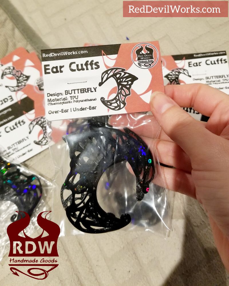 3 designs in black from Red Devil Works new lineup of ear cuffs. Packaged nicely in bags and labels. The designs read: Dragon, Butterfly and Drips. Held up to camera is the butterfly design  