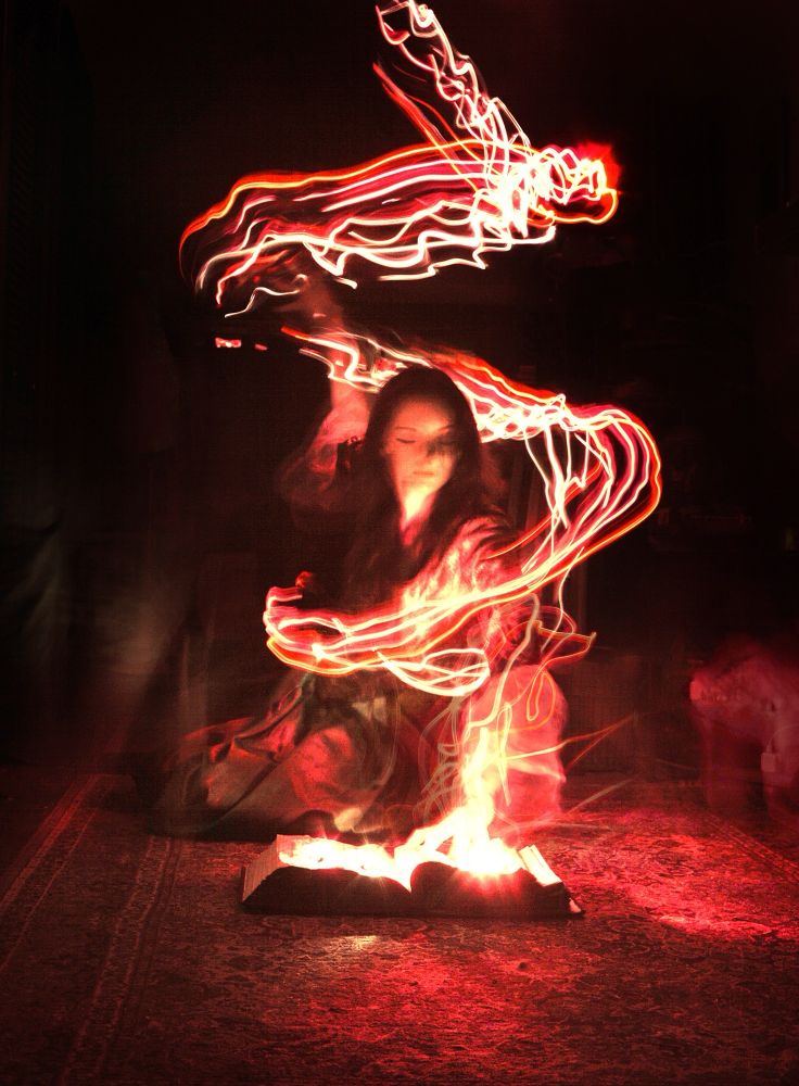 A woman kneels on the ground facing the viewer with one hand on a thick open book.  Out of. The book tons of red light that illuminates the scene pours up her arm then swirls around her and past her other arm out stretched above her head.  The surrounding environment is a dark attic room.