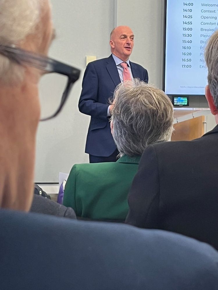A man in a suit stood next to a screen presenting to a group of people.