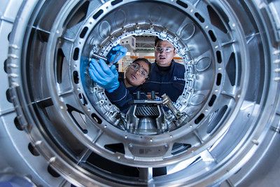 Two engineers wearing goggles, working on and looking through a large piece of metal machinery. 