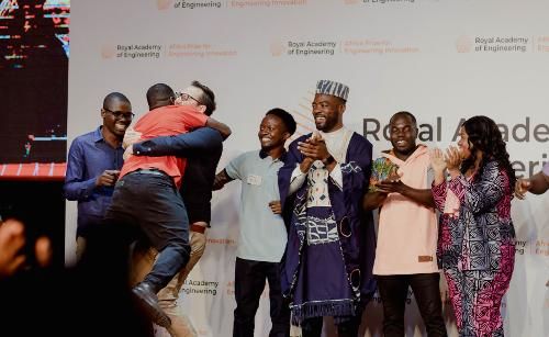 Two men hug on stage, surrounded by five other people clapping and smiling. Behind them, a banner reads 'Royal Academy of Engineering Africa Prize for Engineering Innovation'