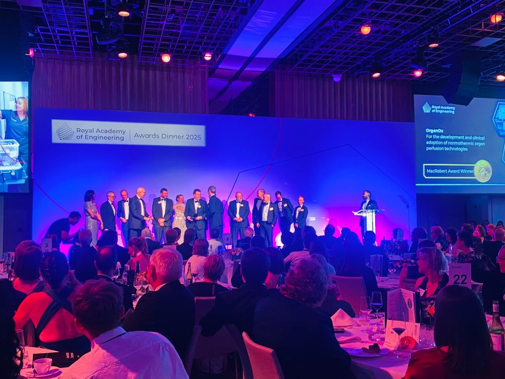 A group of people stand on a colourfully lit stage in a room full of people, who sit at tables and watch them while clapping.