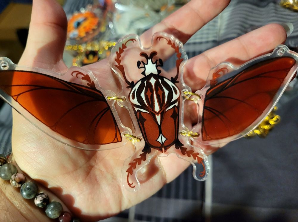 A hand holding a red goliath beetle illustration with spread wings printed on clear acrylic, connected with gold jump rings. 