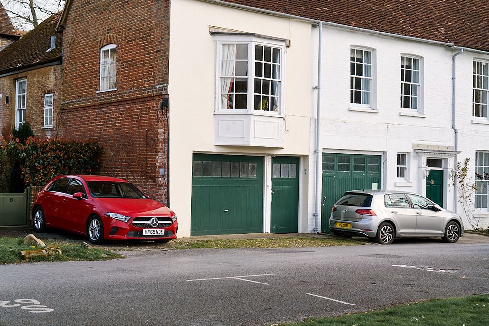 Old mews houses, painted white/cream with dark green doors. Red Mercedes car parked on the left of one house, and silver VW parked at the front of another house. 