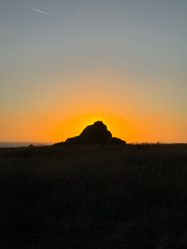 The sun sets behind the rock formation that has a beautiful handmade bench at the top facing the Pacific Ocean at Noyo Headlands Park, Fort Bragg, California. 
