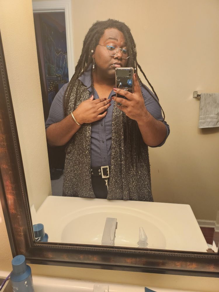 Long locks, tucked in button up blue with the top few buttons unbuttoned and black tee mirror selfie half-body. Red nail polish. Spider bite piercings