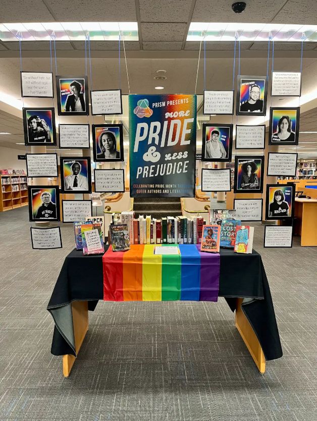 Image of book display sharing photographs and quotes of queer authors hanging above table. The table is draped with a rainbow cloth and shows a collection of our books available.