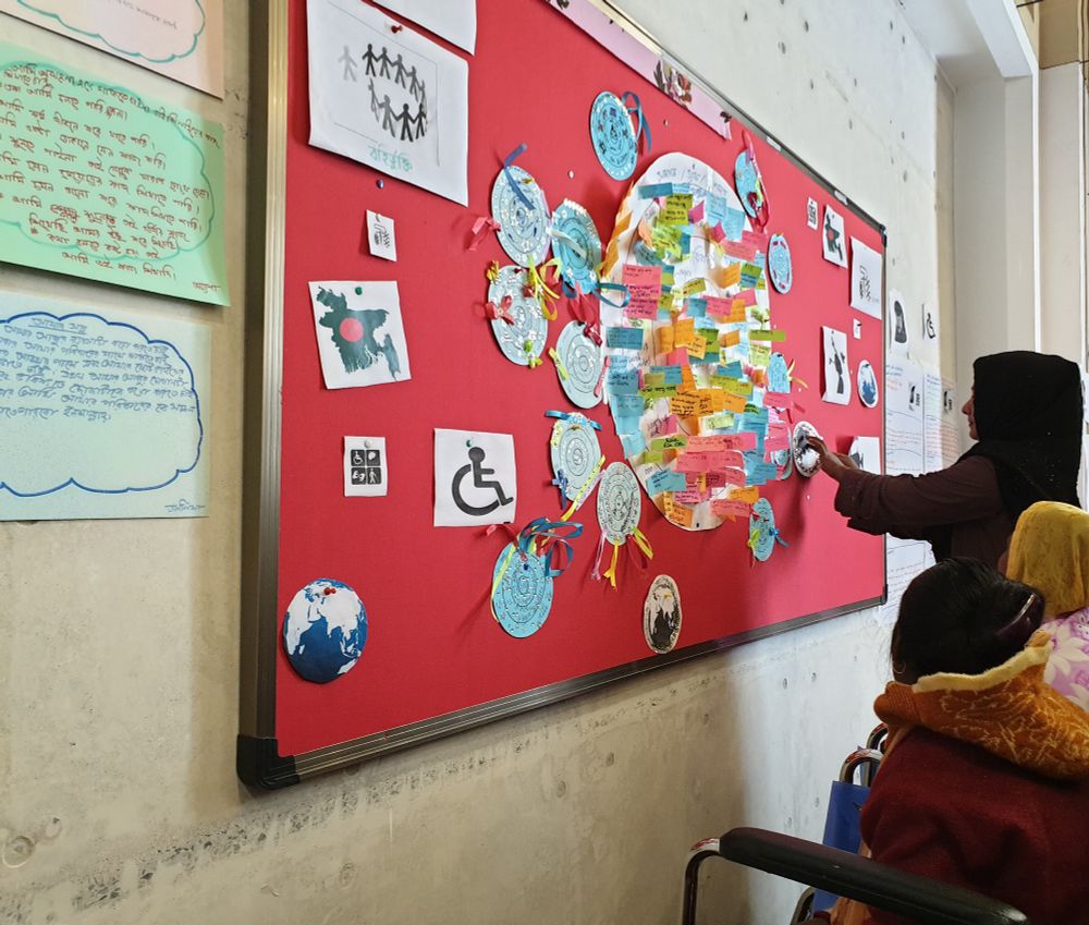 Photo of a group of Bangladeshi women in front of a red noticeboard with lots of pieces of paper pinned to it. 