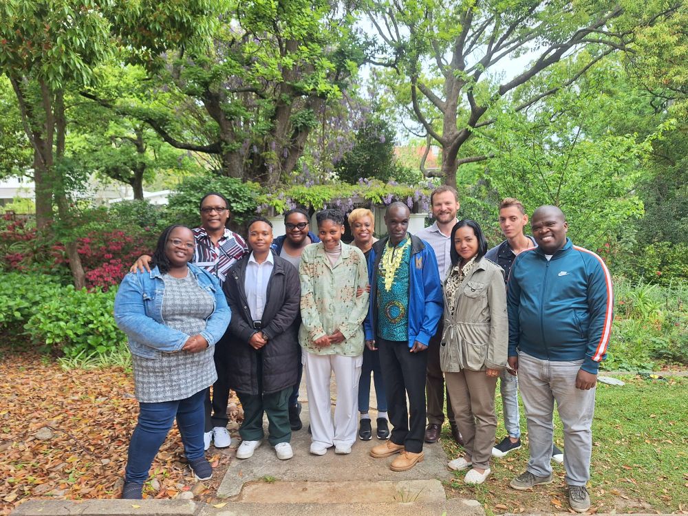 A group of 11 people stood in a garden. They are smiling. There are trees in the background and green plants. 