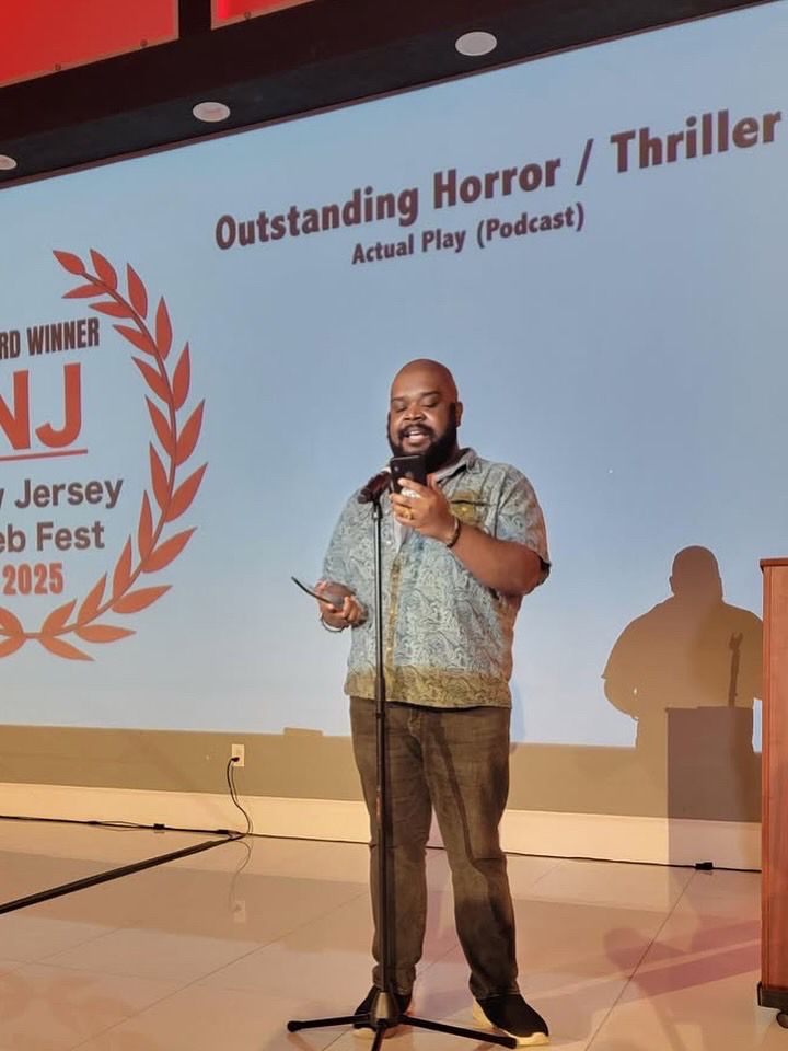 Kendo giving a speech at a mic in front of a screen with the New Jersey Web Fest 2025 emblem and the title “Outstanding Horror/Thriller Actual Play (Podcast)”