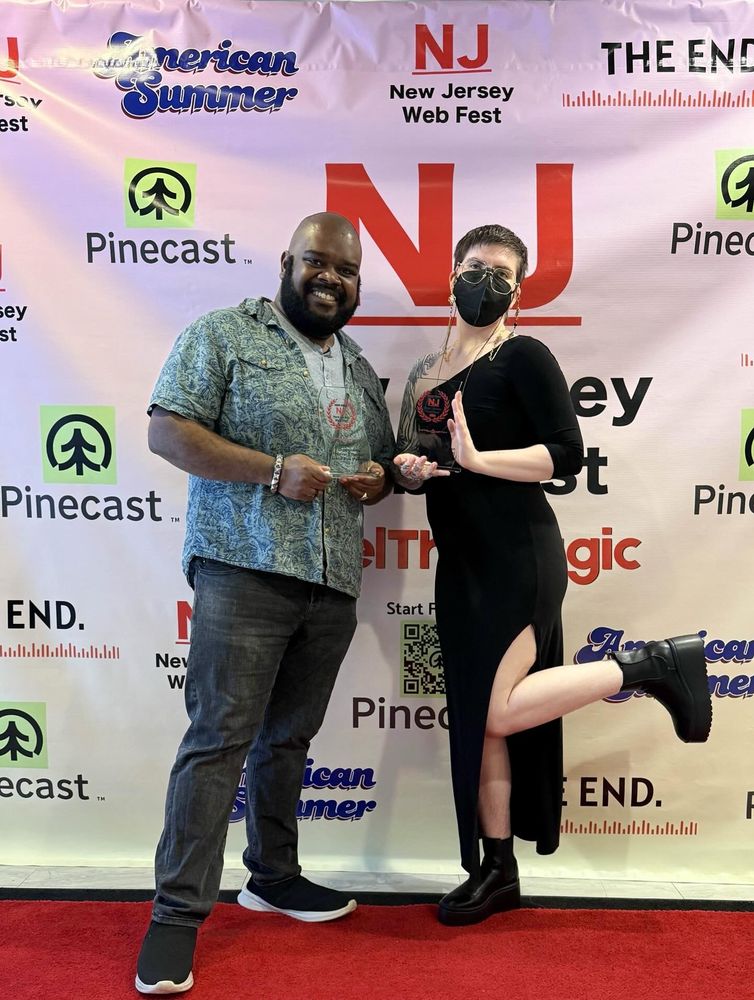 Kendo and Sea holding two glass NJ Web Fest awards while posing on a red carpet with a festival backdrop behind them