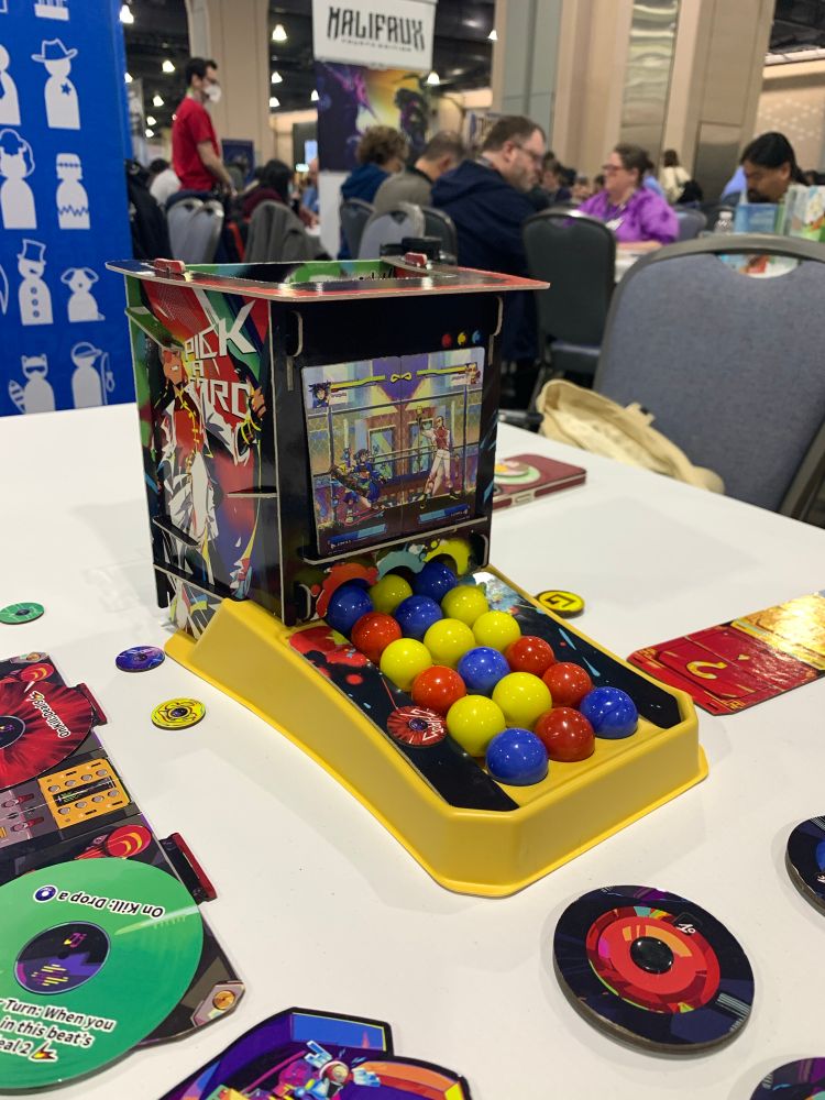 A cardboard arcade cabinet looking boardgame piece with image of a fighting game on it and red, yellow, and blue marbles spilling out from the screen into three different rows. 