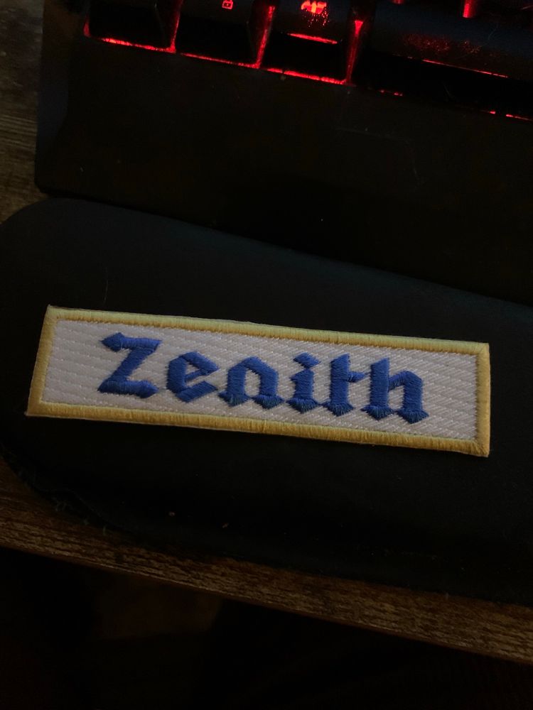 A white, rectangular patch with a gold border. In the center, stitched in blue, is the name Zenith.