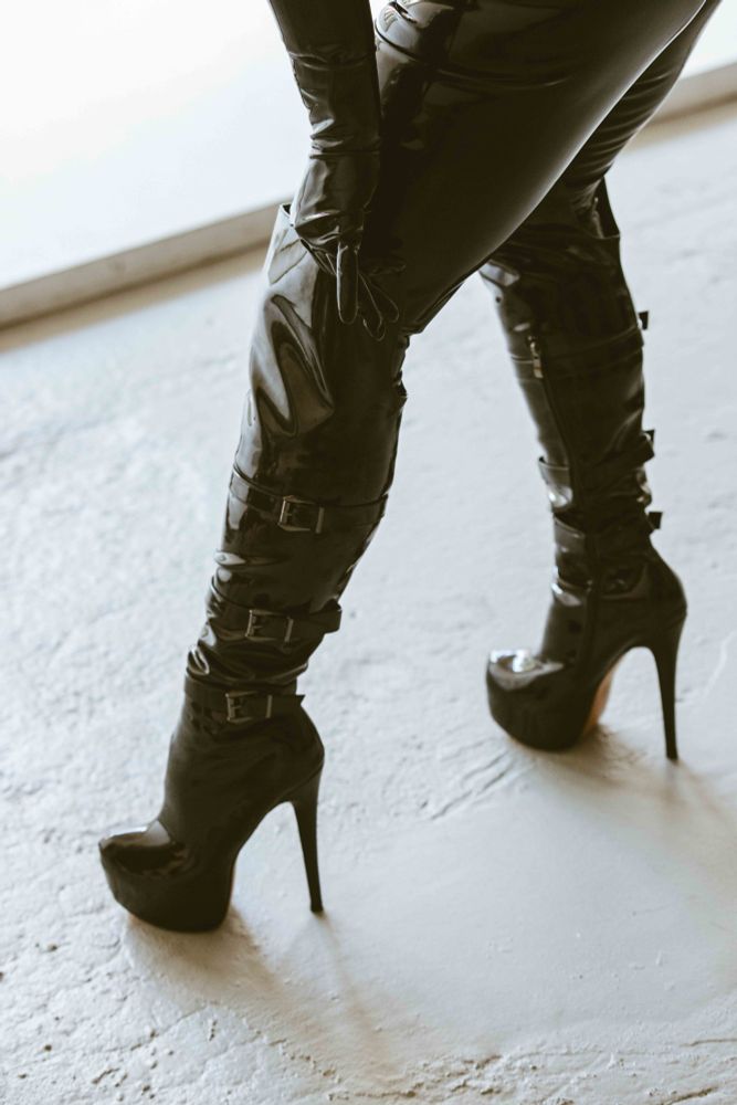 In a bright indirect light, shiny black thigh high stiletto boots with three buckles going up the calf are juxtaposed with the white matte painted surface of the cement floor. One of Her black latex hands reaches down and is grasping the top edge of one shiny boot. One of Her shiny latex curvy thighs is visible above the boot, with a slight hint of shiny ass. 