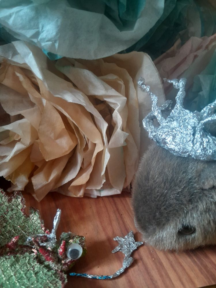 A glittery tiny Dragon in a unicorn horned tinfoil tiara, proffering a star-topped magic wand to a cuddly Wombat in front of coffee filter flowers dyed with natural dyes and left over paint water