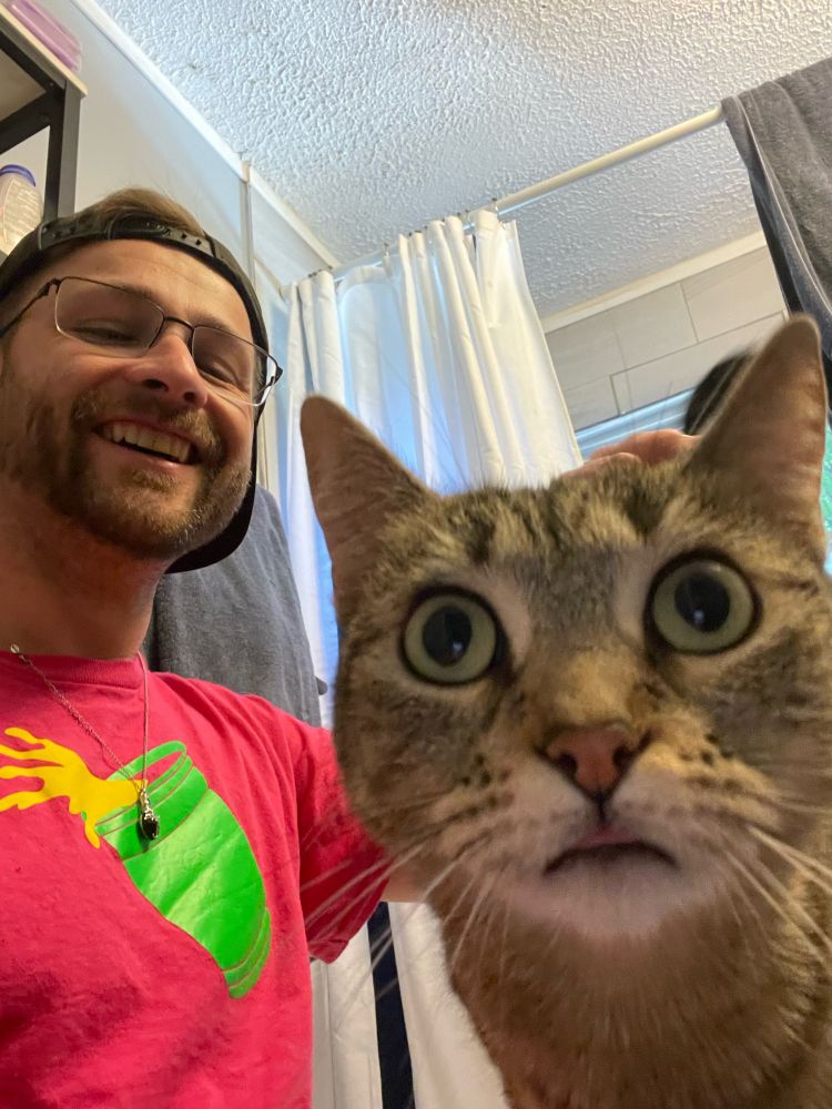White bearded, bespectacled man in pink shirt with green cup iron-on sticker, with backwards cap in background. Gray cat with black stripes and open green eyes in foreground staring at camera. 