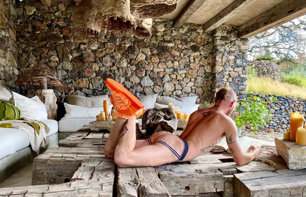 The setting is a stone grotto filled with luxurious white canvas couches. A white man wearing bright orange hi top sneakers and a jockstrap is laying on his stomach on a large coffee table made of wooden beams. He is facing away from camera reading a book with his knees bent and his feet crossed. In the background is a hillside with rock walls that are terraced.