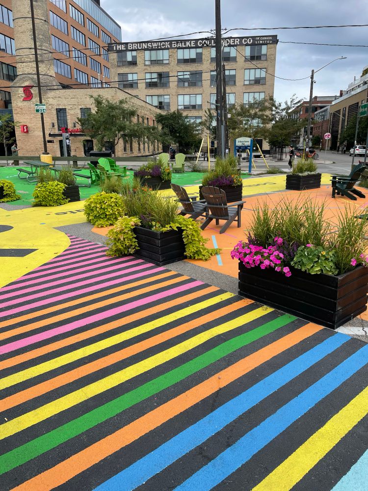 Photo of a parking lot that has been painted bright colours, had planters placed, and chairs & tables placed on it. A temporary park before a more permanent one.