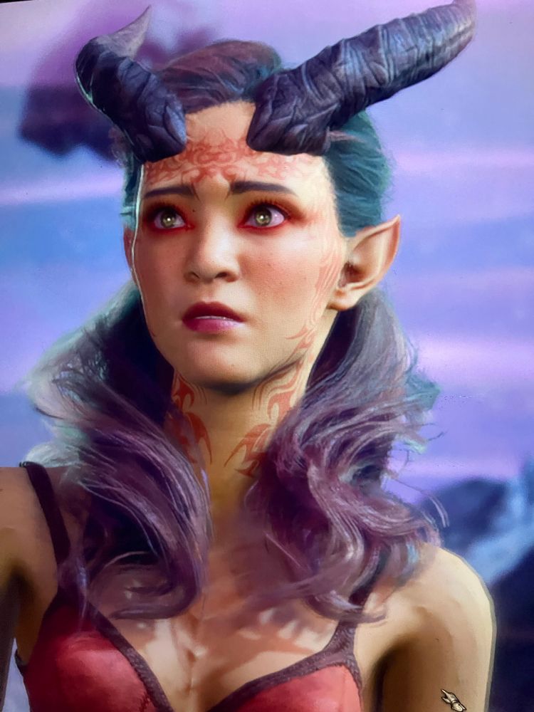 Picture of my character from Baldur’s Gate 3, a tiefling with yellowy skin and orange makeup and tattoos 