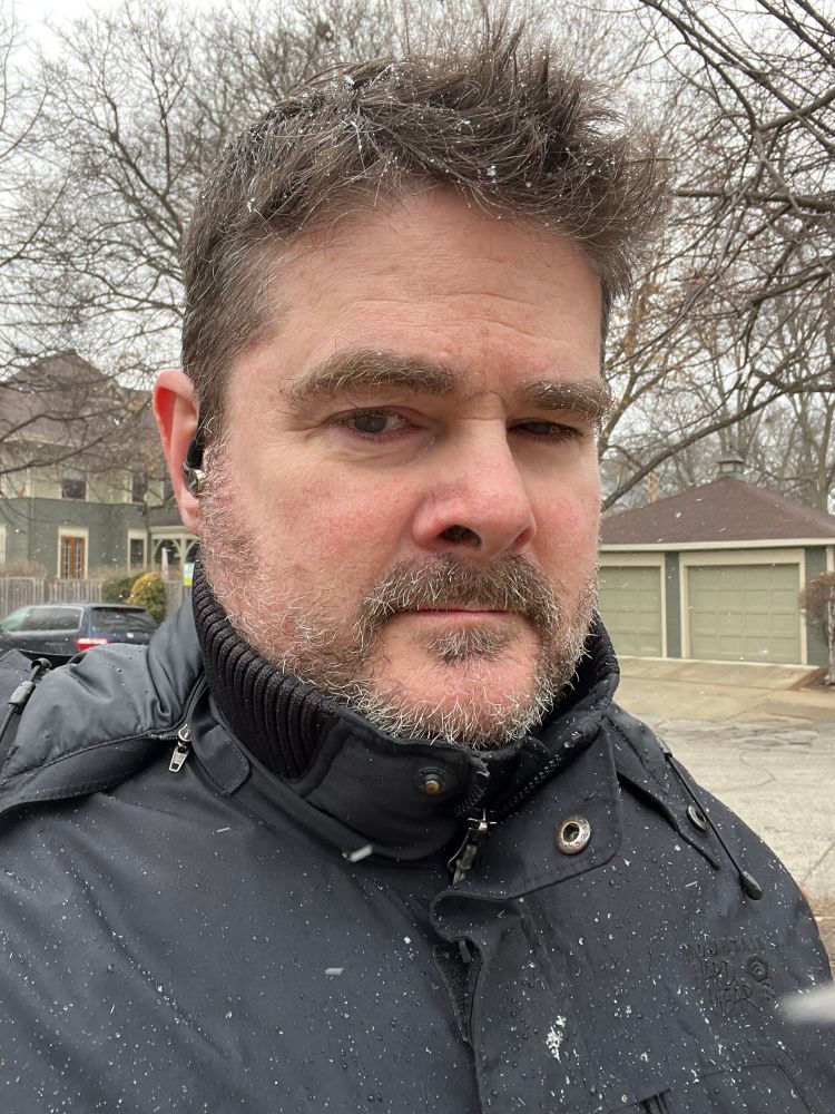 Photo of bearded man with short brown hair. He wears a black winter coat, and it is snowing. 