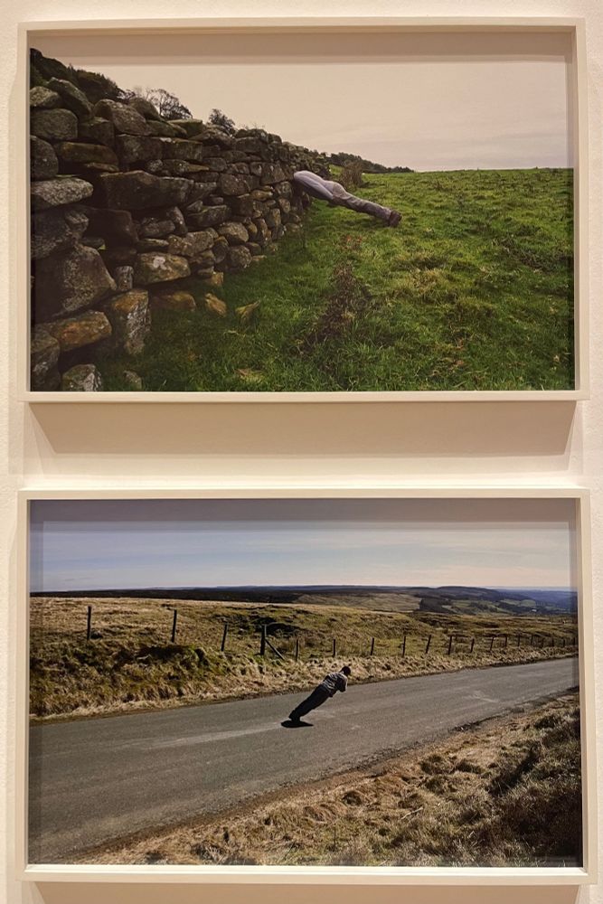 Two photographs, part of a series. 1/ a figure leans at an angle of 30 degrees against a dry stone wall, his head entirely invisible inside it and body rigid as an ironing board . 
2/ A ramrod figure in the middle of a moorland road, leaning at a 45 degree angle, supported only by a strand of barbed wire. 