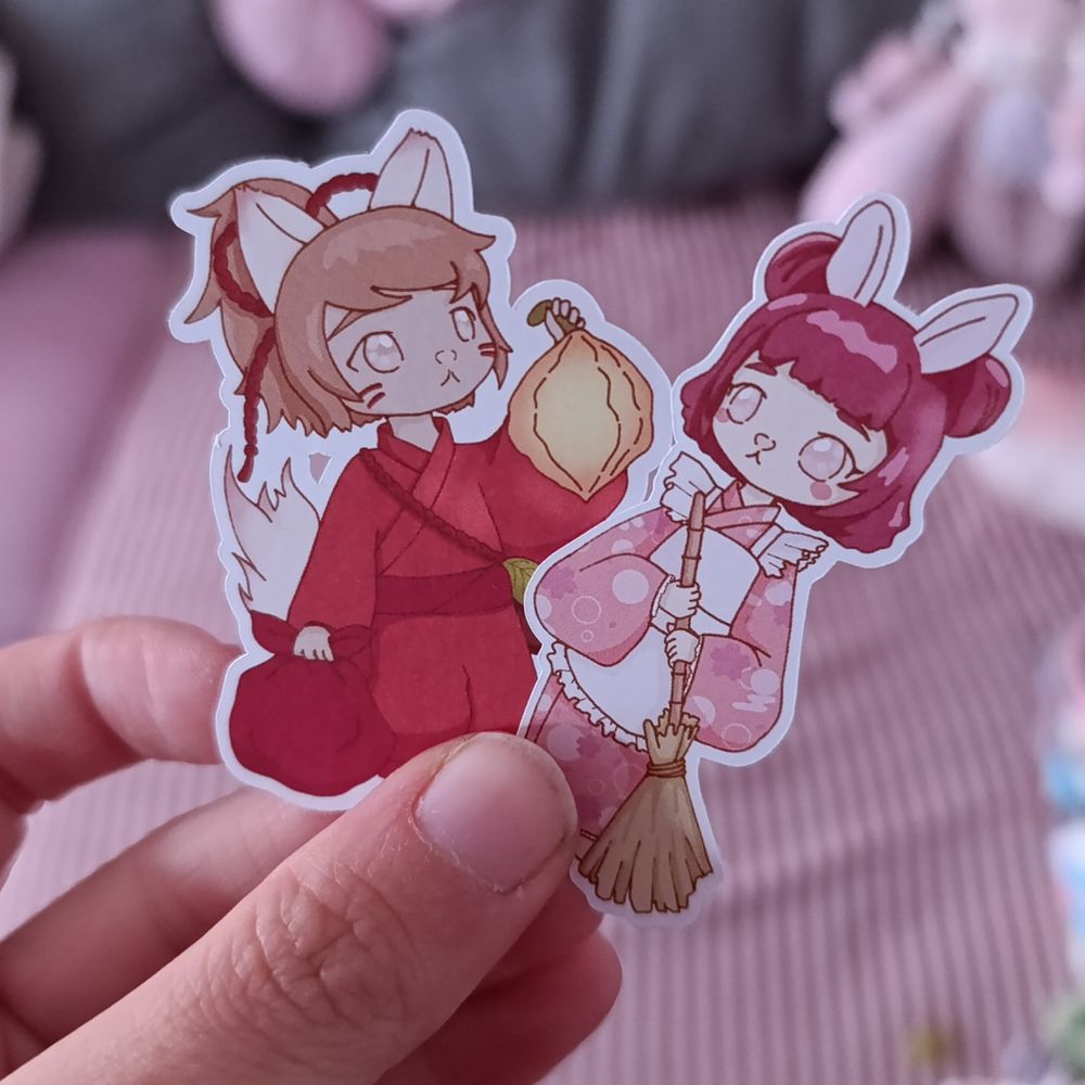 Auf dem Bild werden 2 Sticker in der Hand gehalten. Zwei gezeichnete Charaktere. Einer hat ein japanisches Gewand an mit braunen Haaren, Fuchsohren und einer Physalis als Laterne. Der Zweite hat einen rosa Kimono mit Schürze an, dunkelpinke Haare, einen Besen in der Hand und Hasenohren. 
