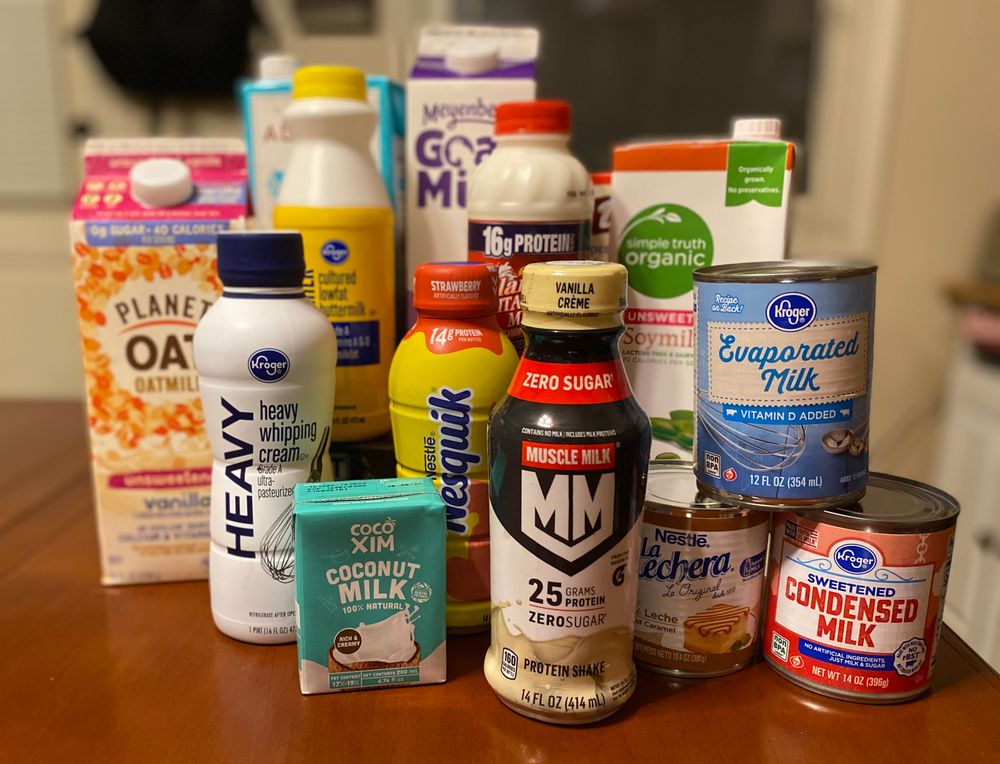 A collage of assorted “milks”, with a box of Betty Crocker cake mix hidden in the back. They include: oat milk, heavy cream, buttermilk, almond milk, goat milk, strawberry milk, whole milk, coconut milk, soy milk, Muscle Milk, sweetened condensed milk, Dulce de leche, and evaporated milk.