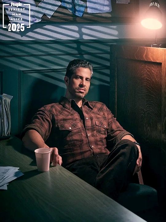 Imagem do ator Adam Rodriguez em uma edição recente da revista People, posando para o photoshoot Sexiest Man Alive 2025. Adam está sentado em uma cadeira de pernas cruzadas, usando uma camiseta quadriculada Isabel Marant e calcas pretas John Varvatos. Sua expressão é séria olhando para a câmera, a barba está por fazer e o cabelo escuro apresenta fios grisalhos. A seu lado, existe uma mesa de madeira escura em cujo tampo ele apoia a mão direita, próxima a um copo branco que aparenta ser de papel.