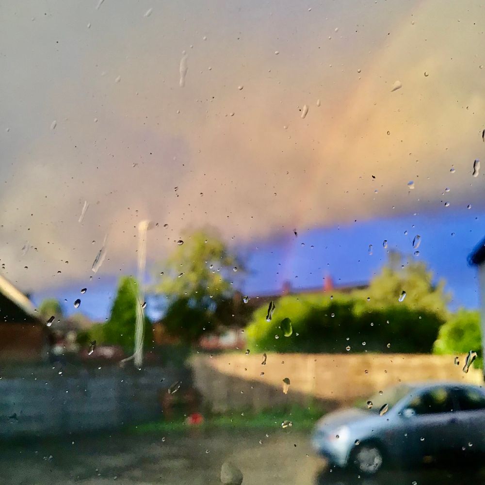 Photo of a rainbow with blue skies and cloud in the background, trees a fence and a car (my Ka) in the foreground, blurred as the camera is focused on raindrops on the window 
