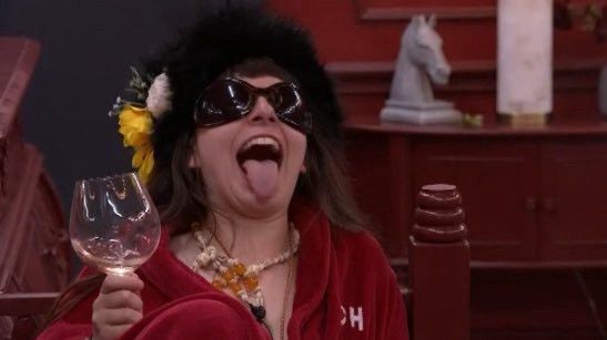 Ava with her tongue out holding a glass of wine in hoh robe wearing her signature hat and sunglasses