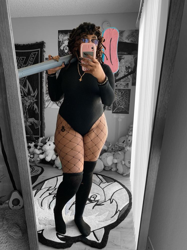 Black woman cosplaying as shizuku from the anime hunter x hunter — she is wearing a black body suit turtleneck, fishnet stockings, black cotton thigh high socks, gold jewelry and is holding a homemade prop from the anime called ‘blinky’ which is a monster vacuum. Also featured a spider with the number 8. 