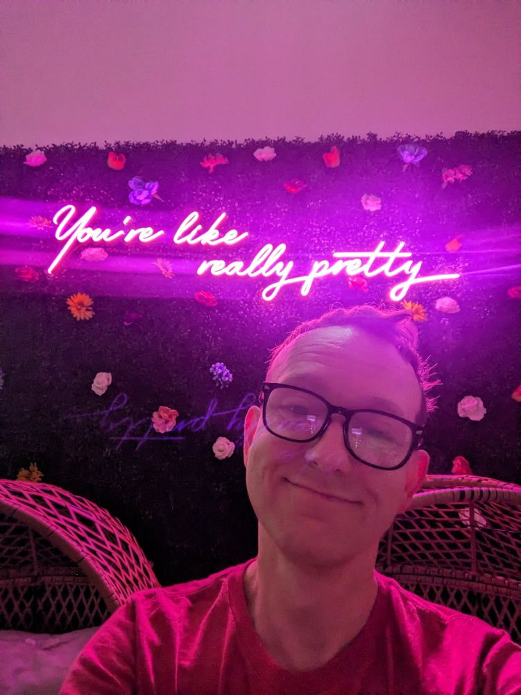 Smiling and wearing glasses in front of a pink neon sign that reads "You're like really pretty" 