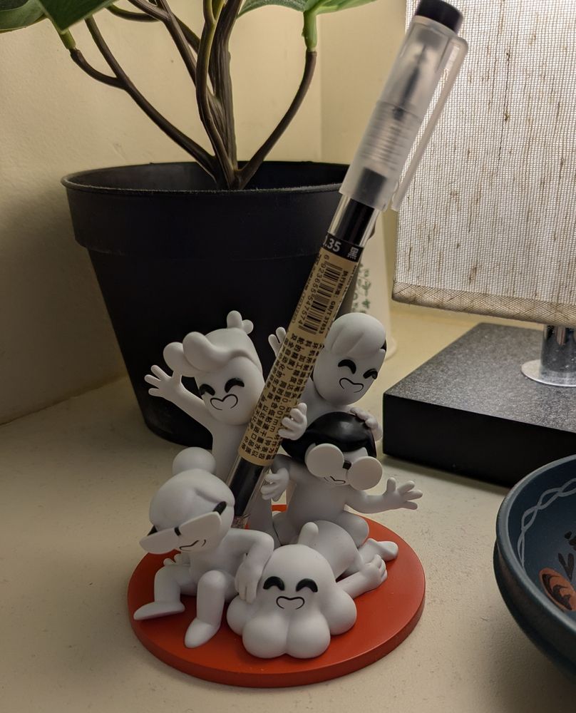 The Drawfee stylus holder (clockwise featuring the Newsdays versions of Jacob, David, Julia, Karina, and Nathan) holding my favorite .38 tip pen. Behind it is a fake plant (windowless office), a lamp, and to the side a small holder dish.