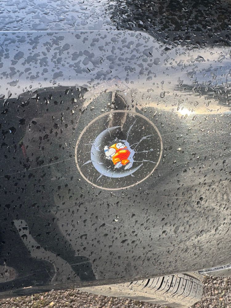 A photo of a transparent sticker of Yamcha from Dragon Ball, dead in a crater, placed in the dent of a car 
