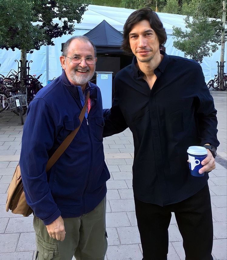Adam standing with an older man/fan (sorry man, I don't know who you are). It daylight and Adam looks like he's still waking up because he's holding a cup of coffee and his eyes are adorably sleepy looking. Adam's wearing a dark blue shirt, black pants, has a bit of stubble, and he's smiling a little with one hand on the man/fan's shoulder.
