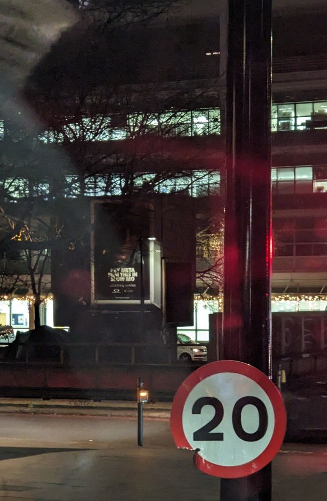 Night time scene in London with speed limit sign for 20 miles an hour in foreground and an advert saying Hey Meta. Film this in slow mo behind 