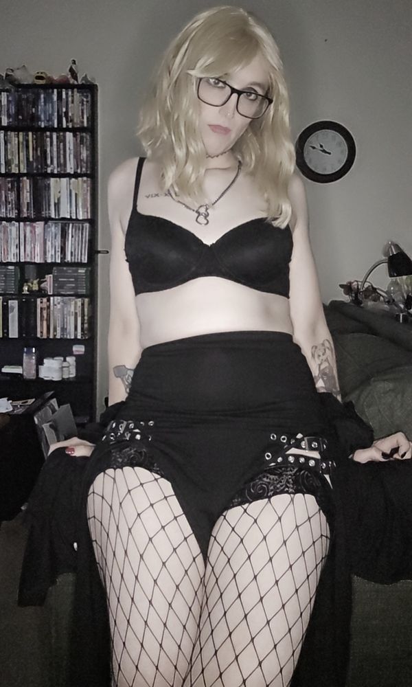 Blonde milf in a black bra, buckled skirt, and fishnet thigh highs, leaned up against the arm of a couch.