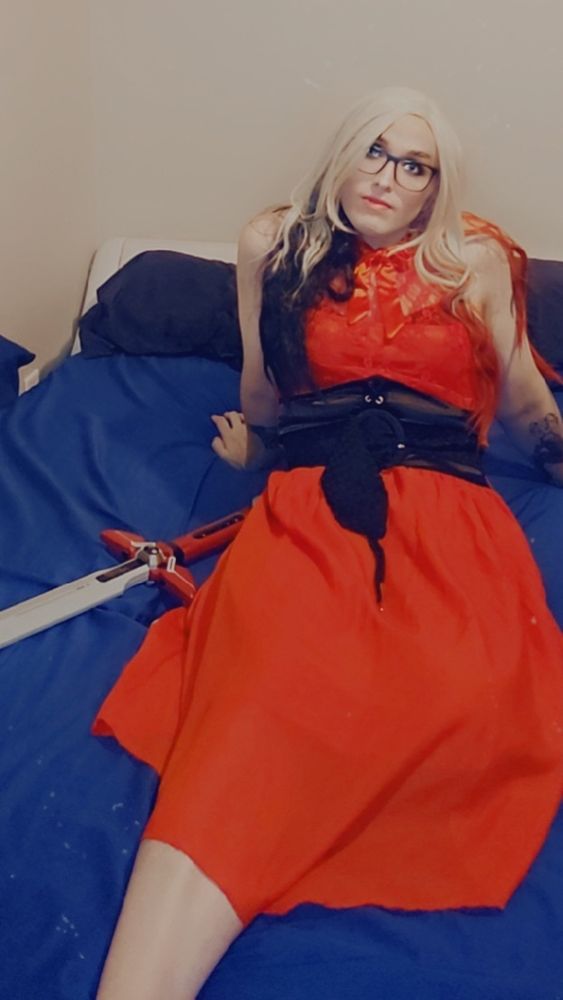 Blonde girl sitting on her unmade bed in a red dress. She has a nerf sword laying next to her because she's obviously super cool.
