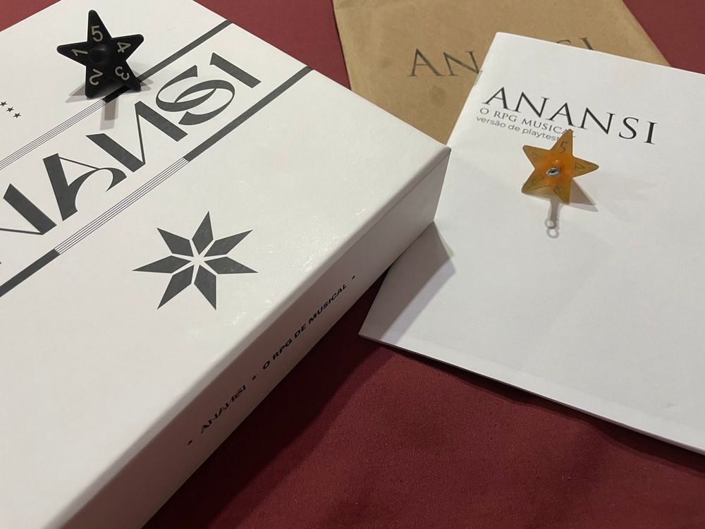Caixa do Anansi com o dado de estrela em cima, ao lado da versão playtest impressa e protótipo do dado em forma de estrela. 